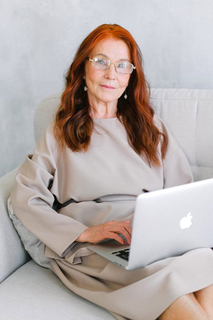Cheerful senior woman entrepreneur with long red hair in dress sitting on comfortable sofa and browsing laptop at home